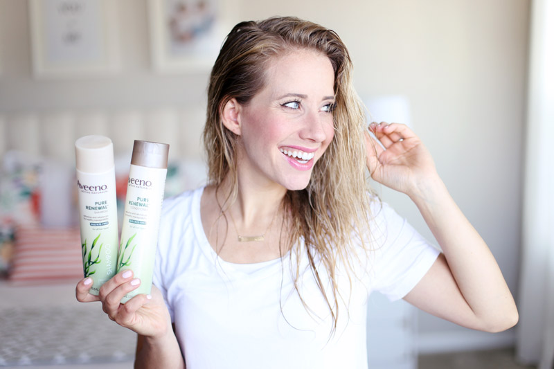 Twist Me Pretty's Abby has wet hair. She smiles and holds bottles of Aveeno Pure Renewal shampoo and conditioner in one hand and touches her hair with the other.