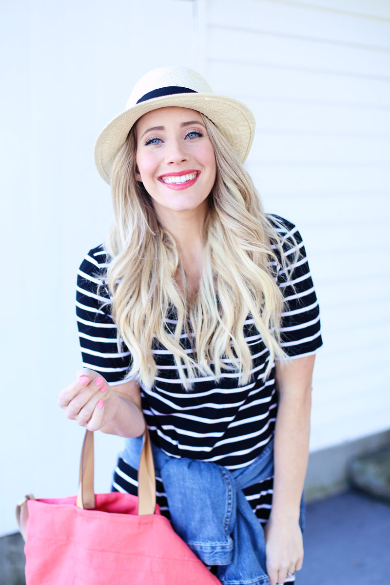 Holding a handbag and wearing a striped blue and white shirt and a wicker hat, Abby smiles at the camera as her hair cascades loosely down her shoulders.