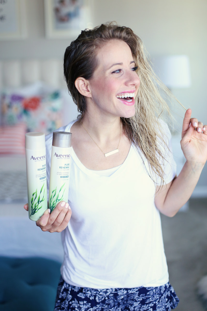 Twist Me Pretty's Abby has wet hair. She smiles and holds two bottles of Aveeno Pure Renewal.