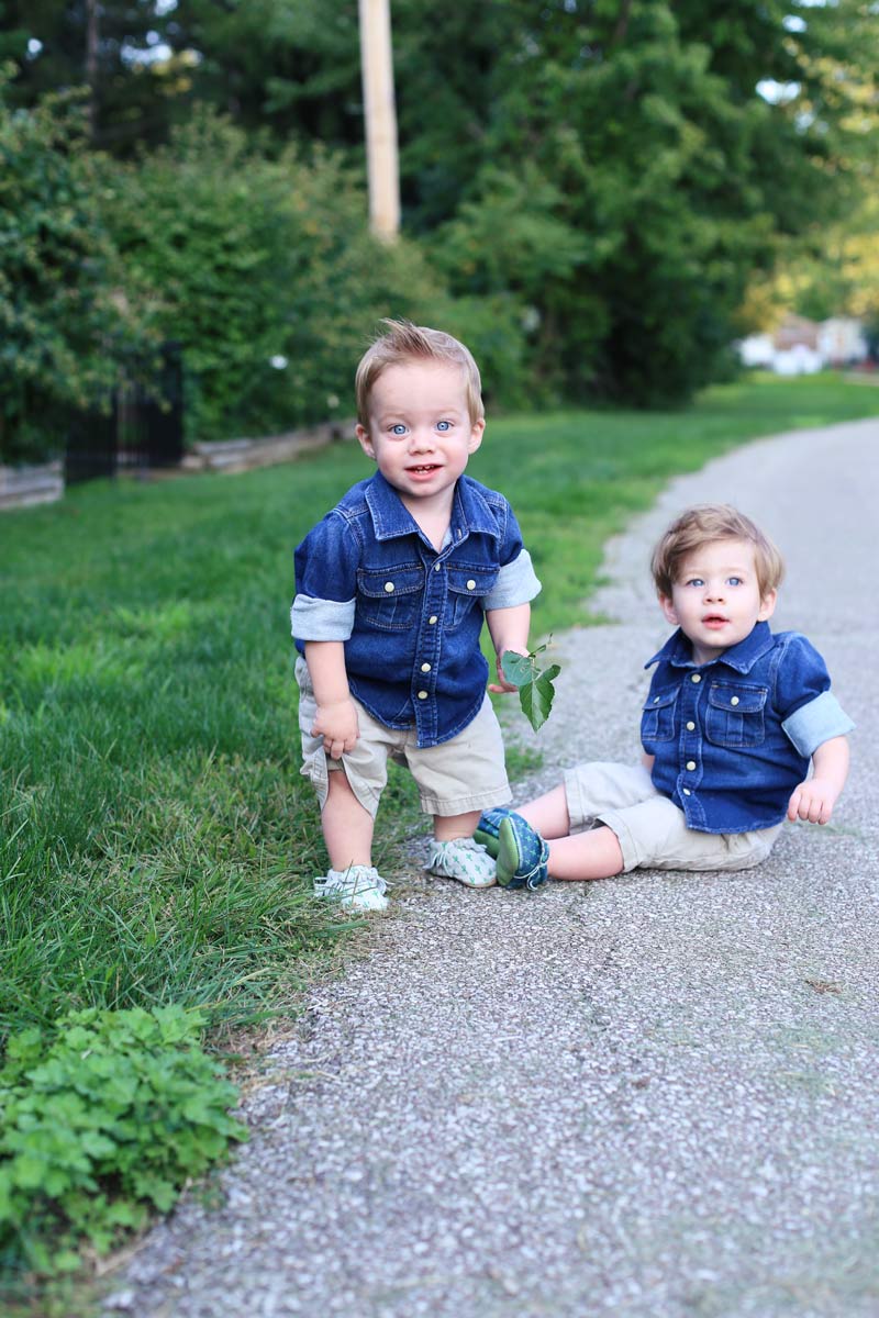 Twin boys with bright blue eyes are wearing denim jackets. One sits, one stands in the park. Enjoy motherhood. Twist Me Pretty.