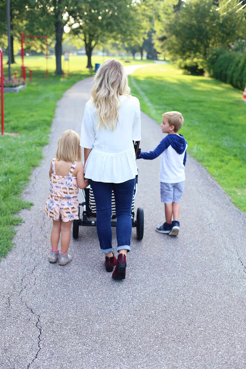 A mother walks away from the camera, pushing a pram, with her daughter on her left and her son on her right. Twist Me Pretty Enjoy Motherhood.