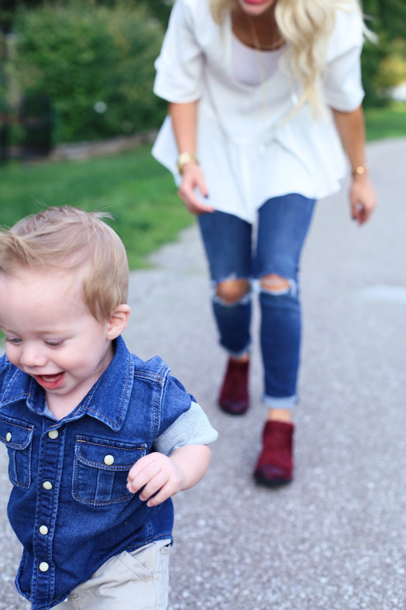 In the foreground, a baby boy runs, grinning. In the background, his mother bends slightly forward as she chases him. Enjoy Motherhood. Twist Me Pretty.