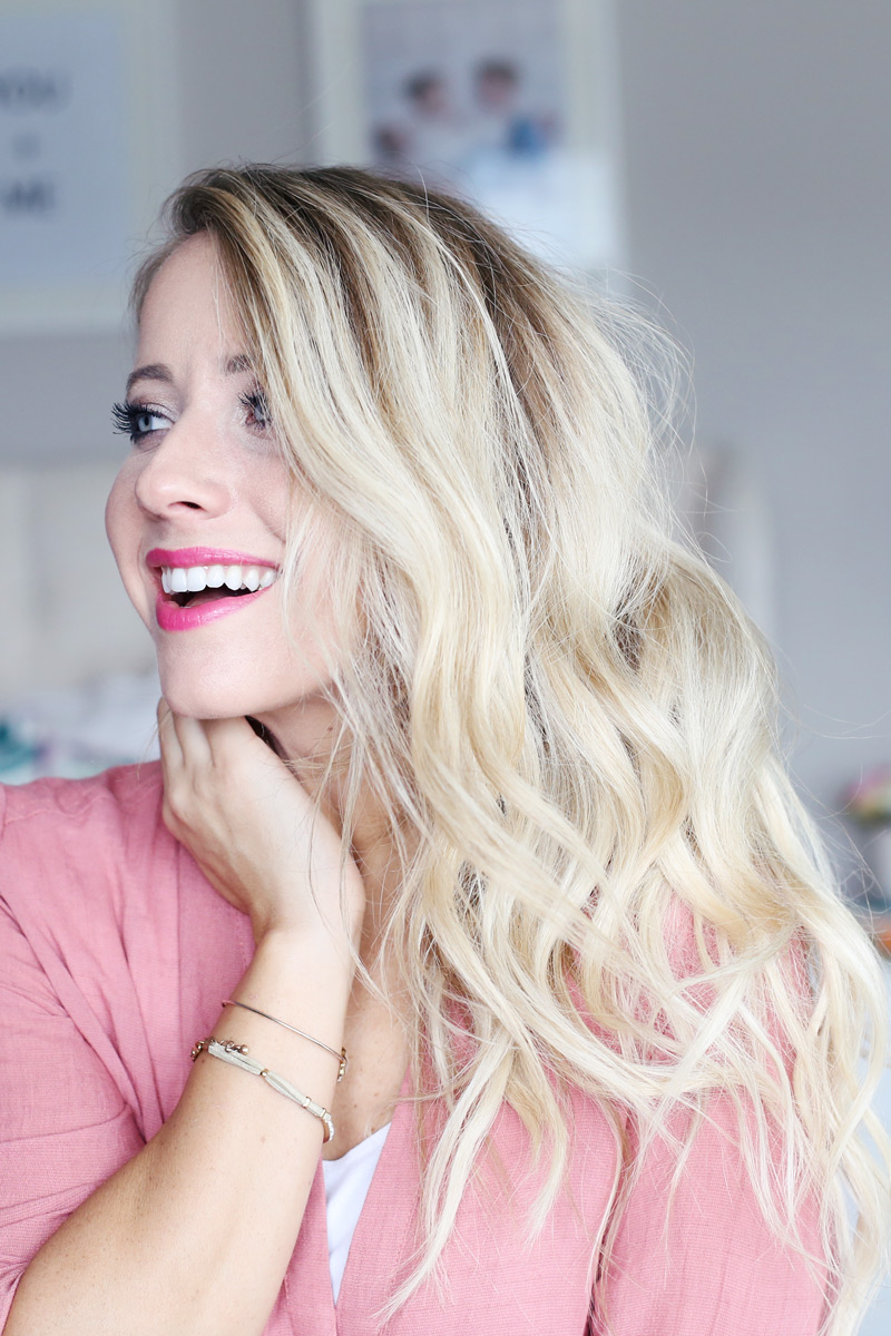 A woman wearing pink lipstick with wavy blonde hair smiles as she looks to her right. Twist Me Pretty. Find how how you can get her gorgeous hair.