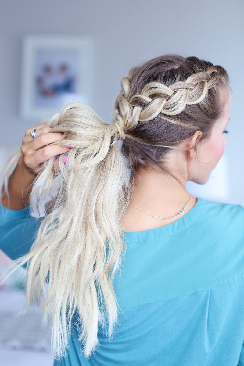 A blonde woman faces away from the camera, holding her hair, which is twisted into a dutch braid. Hair style tutorial.