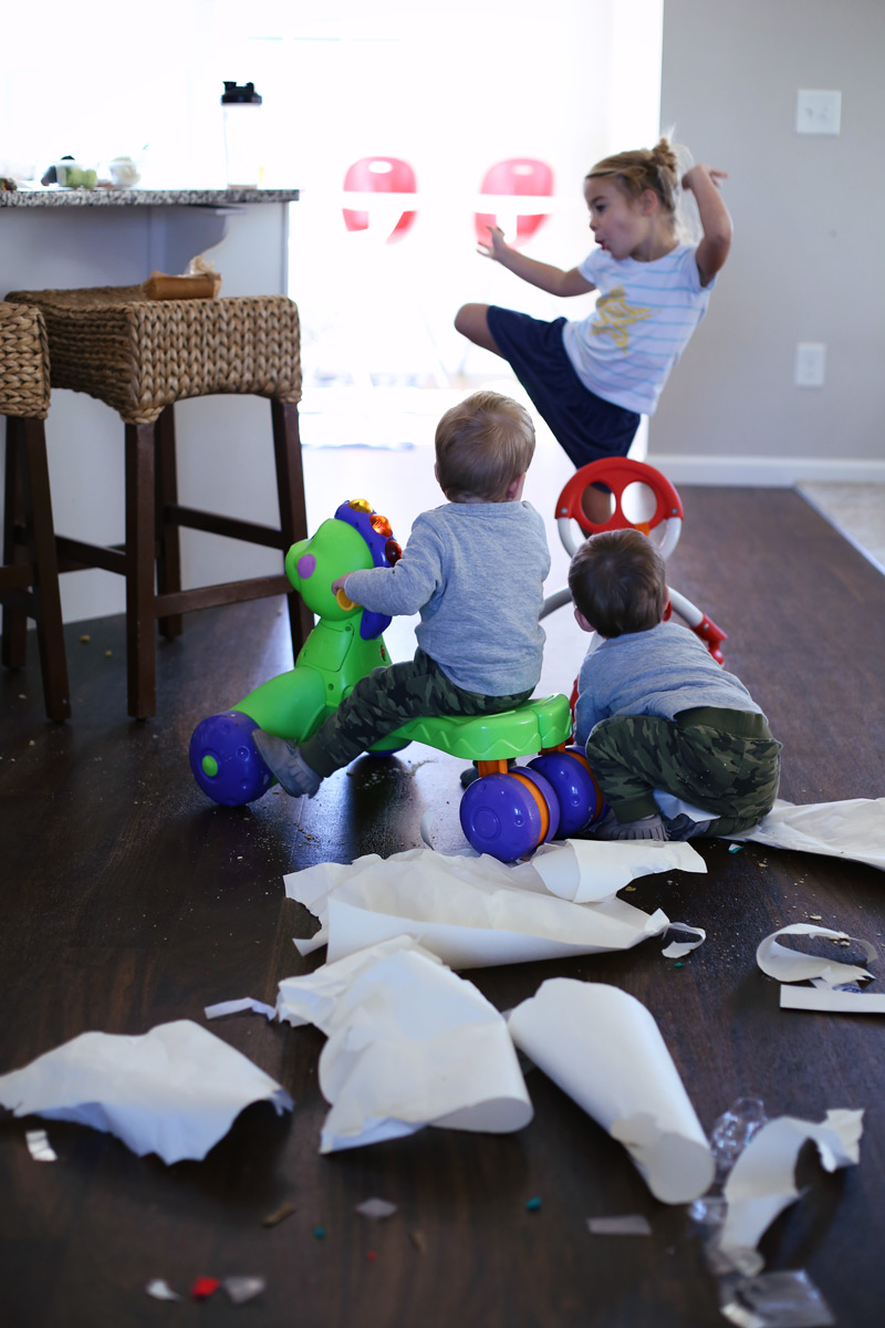 In a kitchen, two toddlers watch their older sister dance, with shredded paper in the foreground.