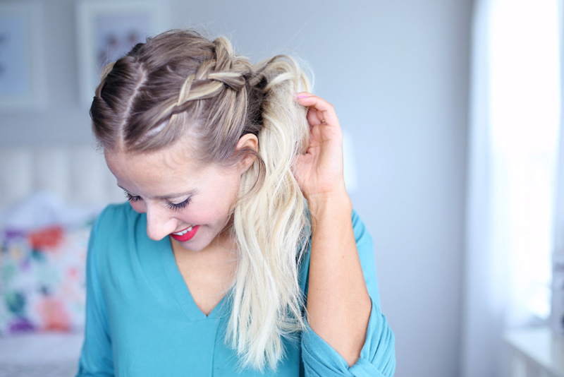 A blonde woman looks down as she holds her hair, which is styled in a dutch braid. Dutch braid hairstyles tutorial.