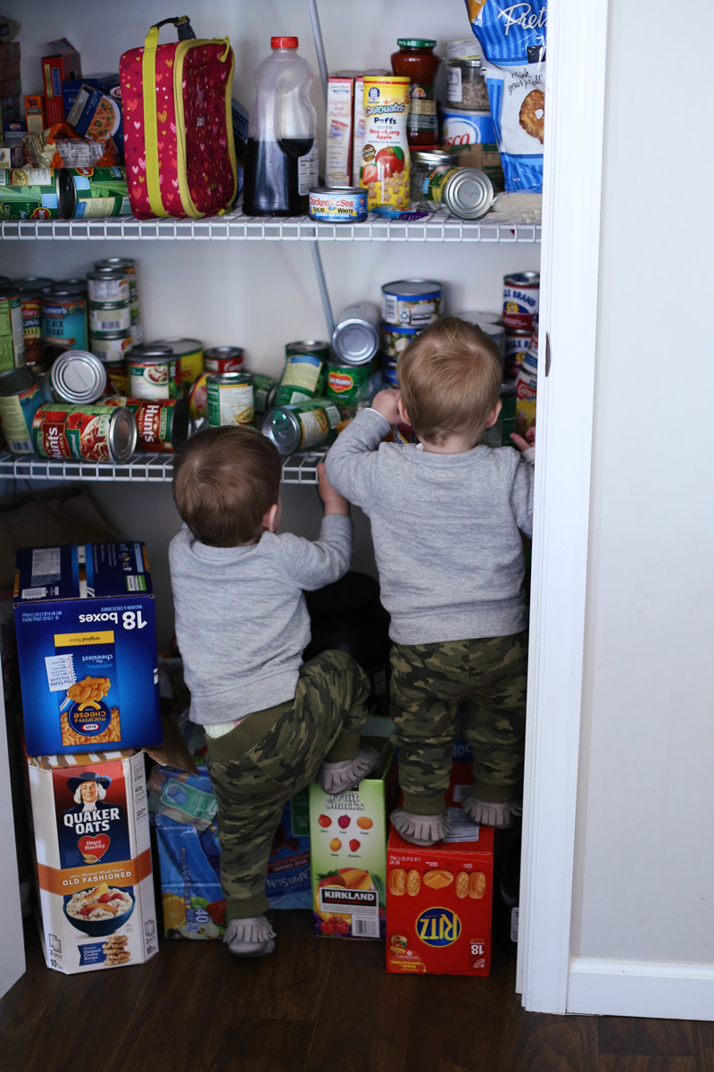 Two toddlers climb on boxes to explore shelves of tins and food.