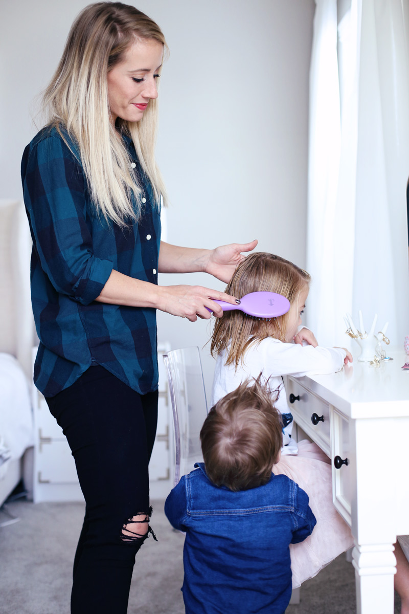 In a light room, a mother smiles as she brushes her daughter's hair while her son watches.