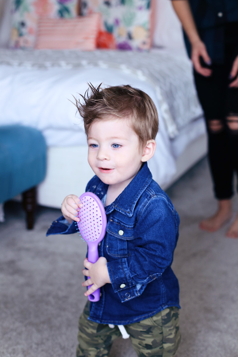 A little boy with bright blue eyes and a cowlick hair style smiles and holds a purple hair brush. Twist Me Pretty - Our Morning Routine.