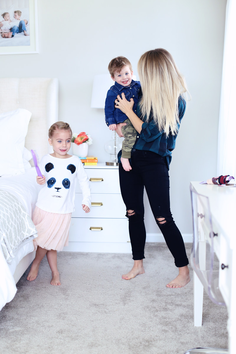 In a bedroom, a mother smiles at her son as he sits on her hip, while her daughter dances with a hairbrush. Twist Me Pretty - Our Morning Routine.