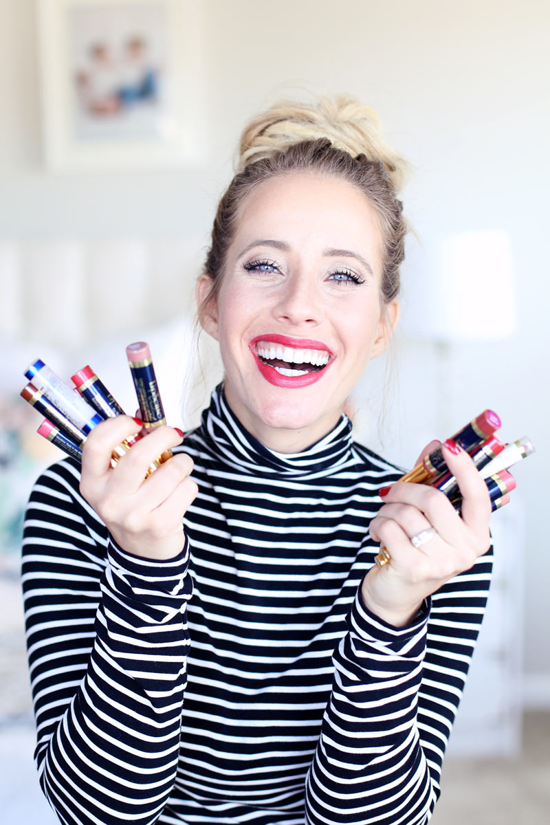 A blonde woman with her hair in bun and wearing a black and white striped polo neck smiles as she holds up two handfuls of longlasting lipsticks. Twist Me Pretty