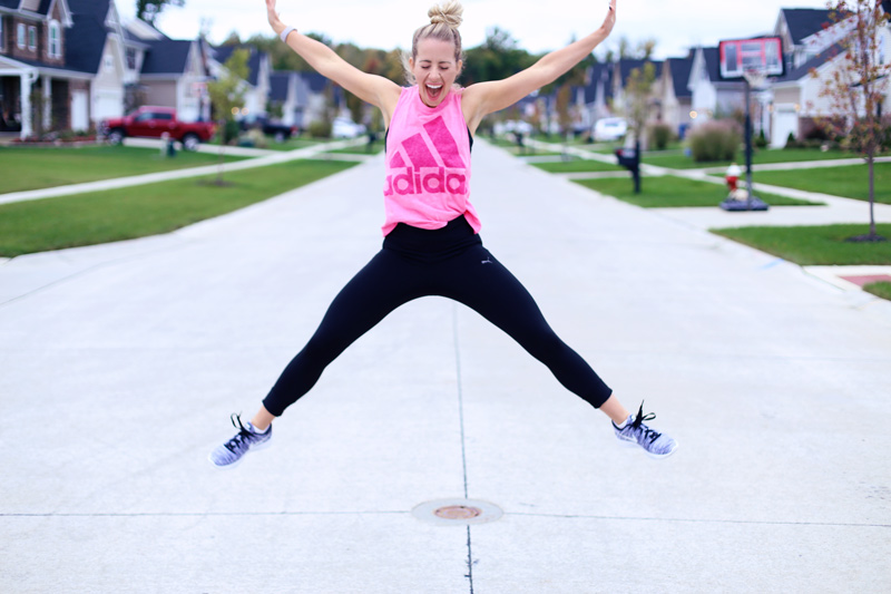 Jumping in the air in a star shape, a woman in sports clothes opens her mouth wide. Doing jumping jacks helps her stay in shape. Twist Me Pretty.